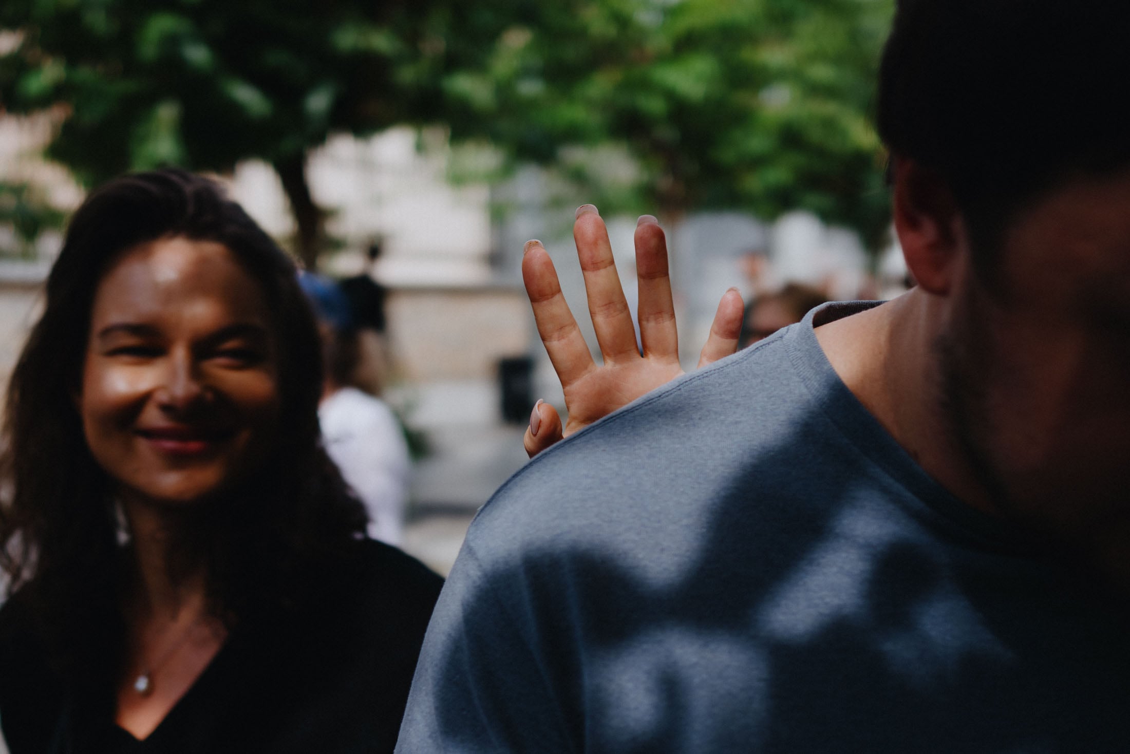 A woman smiles playfully in the foreground while a hand reaches out from behind her to touch a man's shoulder in a sunny outdoor setting.
