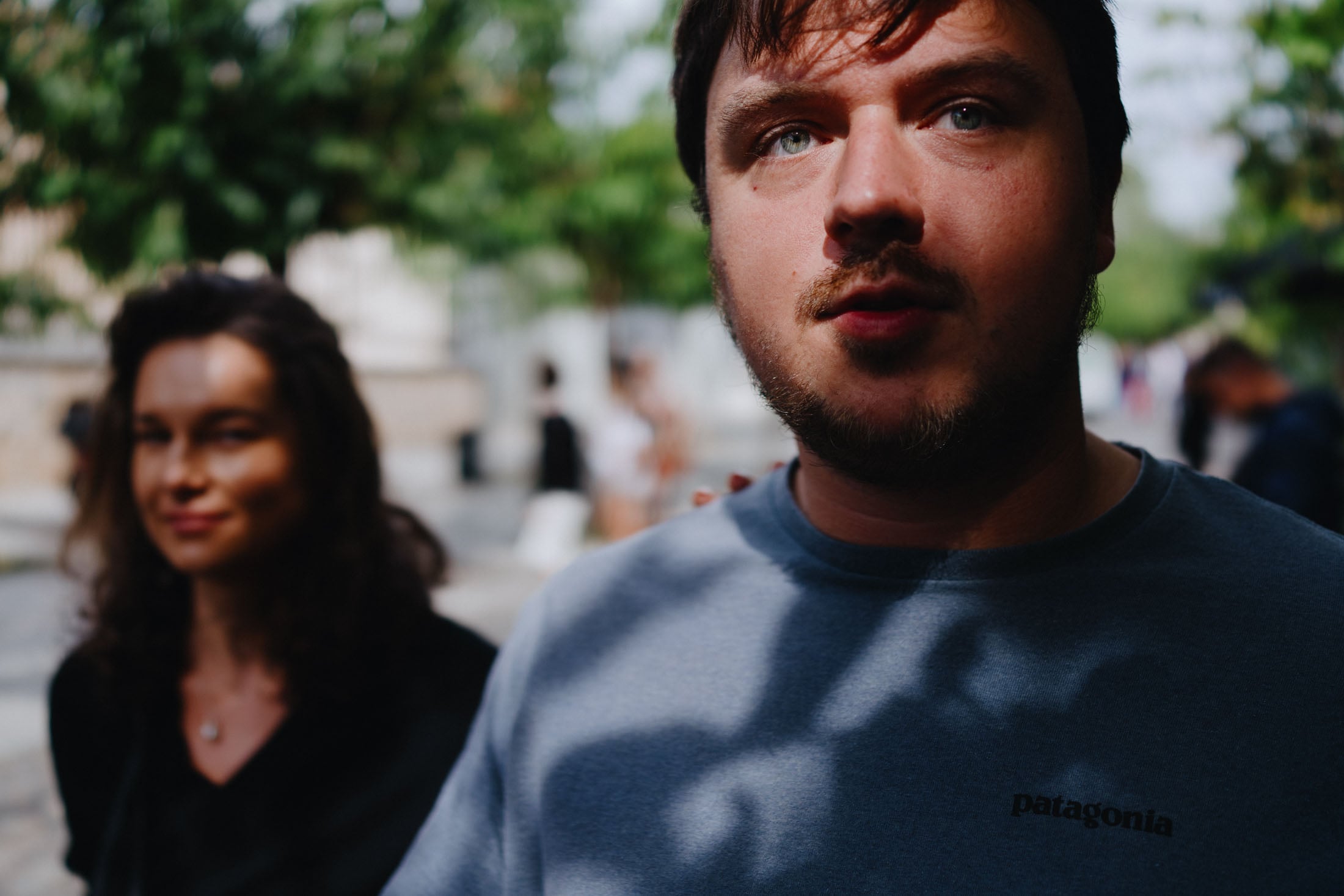 A close-up image of a man with a beard wearing a blue Patagonia sweatshirt, with a woman in a black top slightly blurred in the background. Sunlight creates shadows across the man's shirt, and trees are visible in the background.