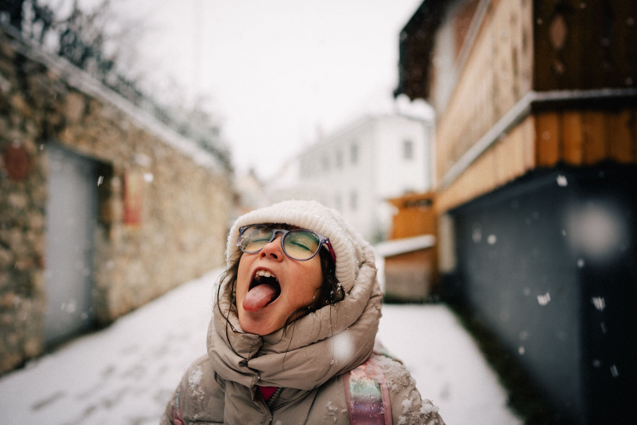 A child with glasses and a winter coat stands in a snowy alley, joyfully sticking out her tongue to catch snowflakes falling around her.