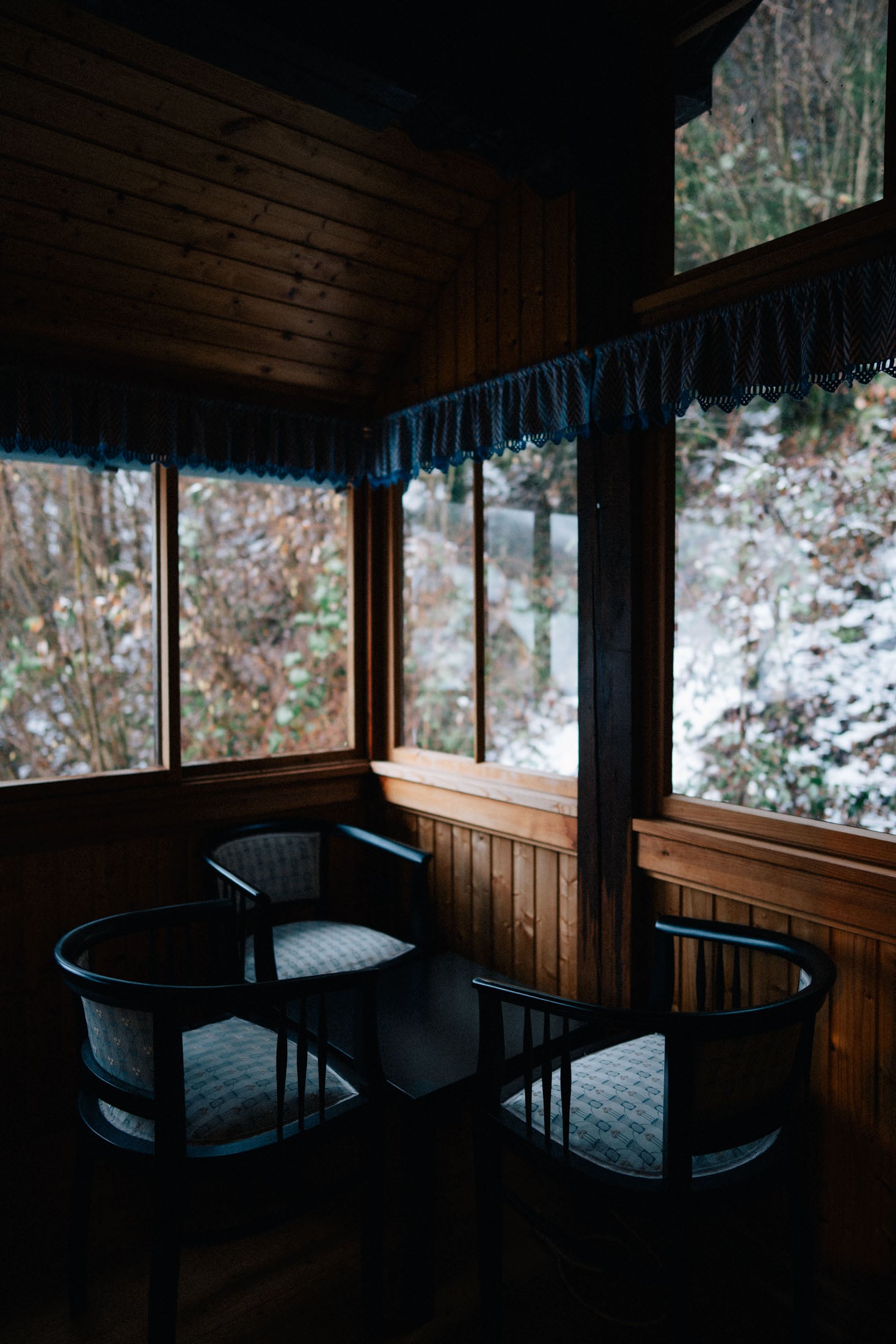 Cozy interior of a wooden room with two black chairs and a small table, surrounded by large windows showing a snowy landscape outside.
