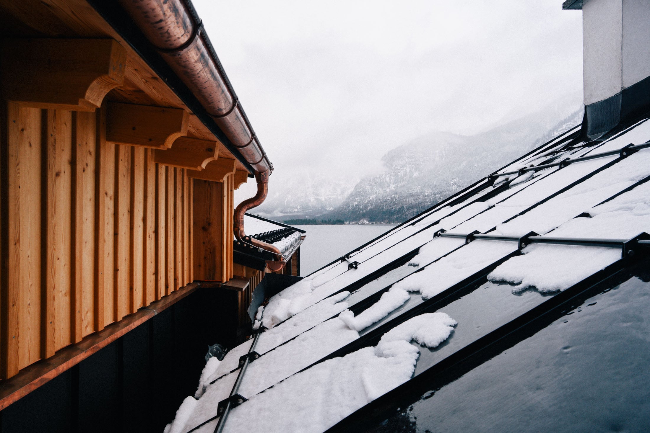 Close-up view of two rooftops, one wooden and the other metal, with snow on the metal roof and a misty mountainous landscape in the background.