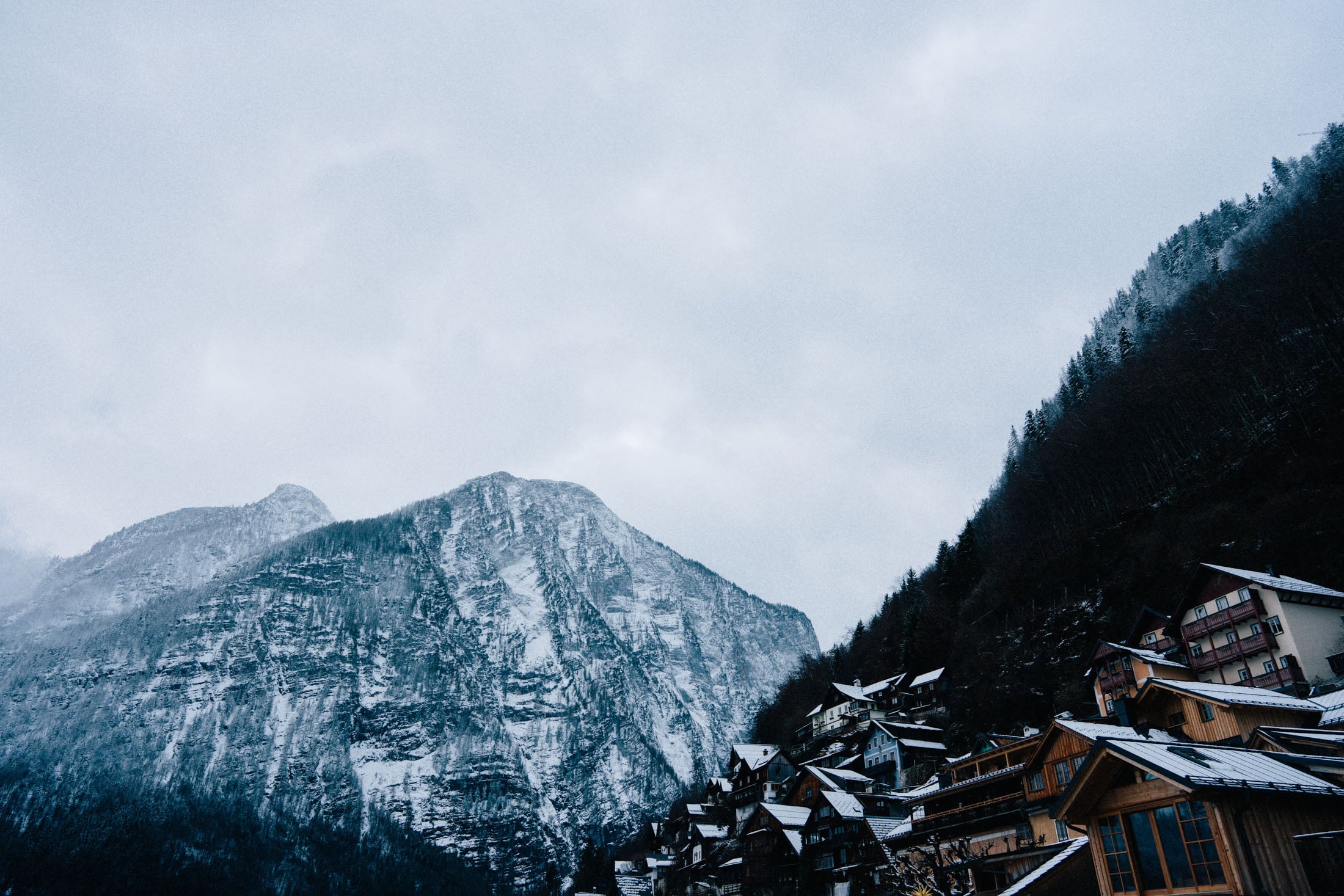 Snow-covered mountains loom over a quaint village with wooden houses, set against a cloudy sky.