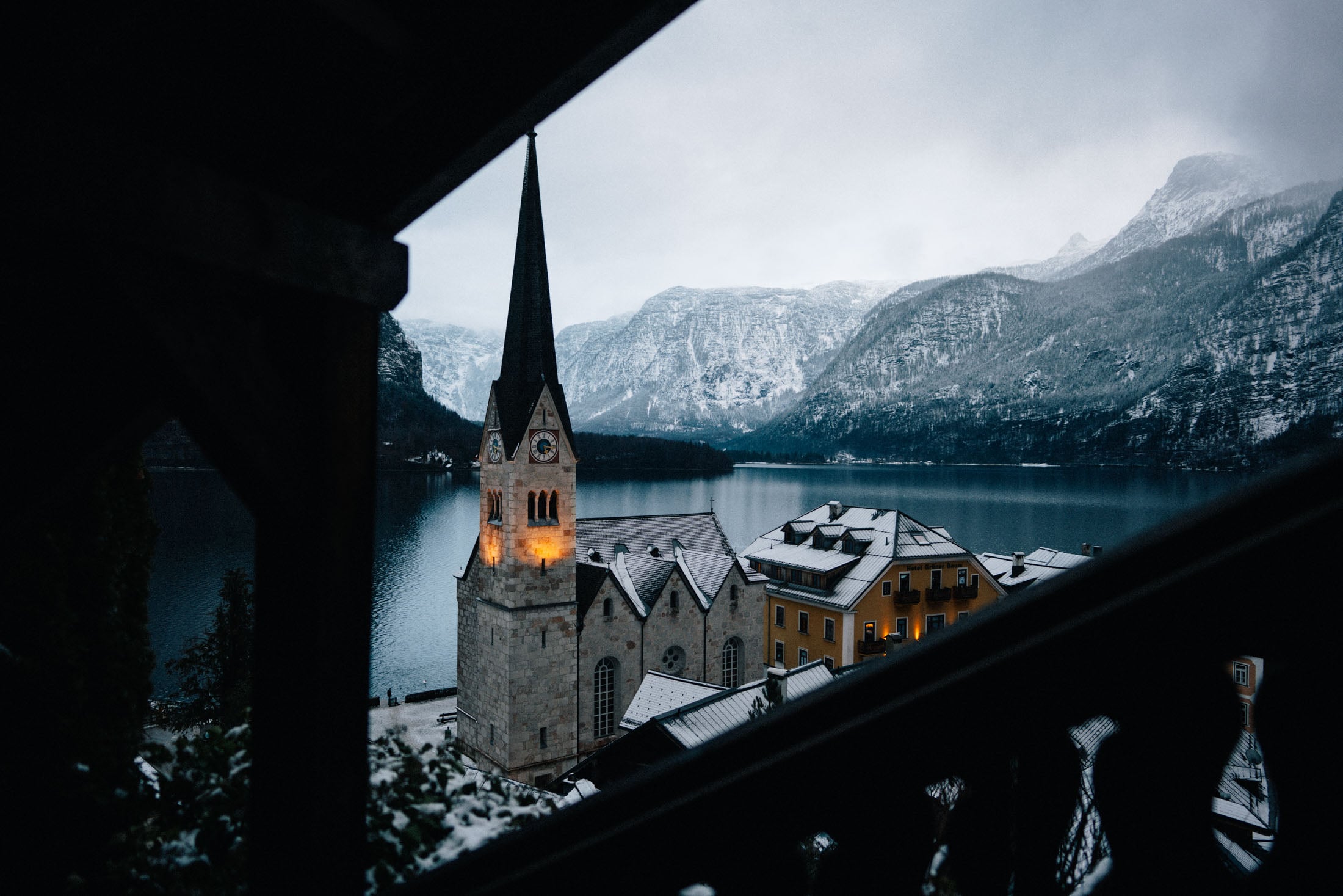 View of a snow-covered church with a tall spire and surrounding buildings next to a lake, set against a backdrop of mountains under a cloudy sky.