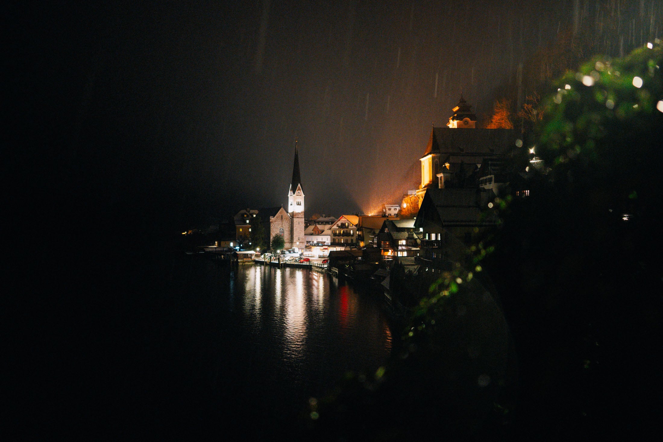 A nighttime view of a serene village by a lake, featuring illuminated buildings and a church steeple, with reflections on the water and a misty atmosphere.