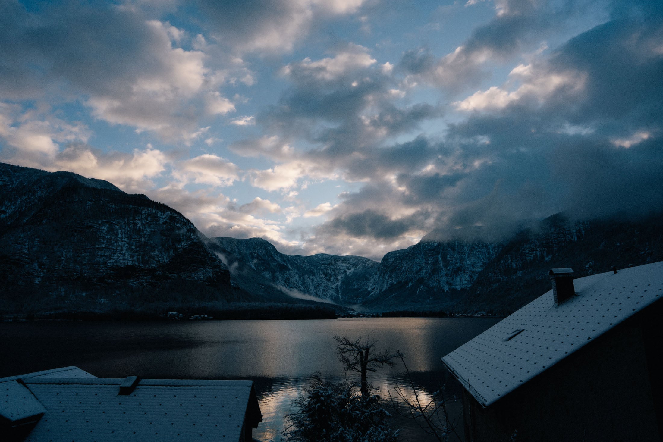 A scenic view of a tranquil lake surrounded by mountains under a cloudy sky, with a hint of snow on nearby rooftops.