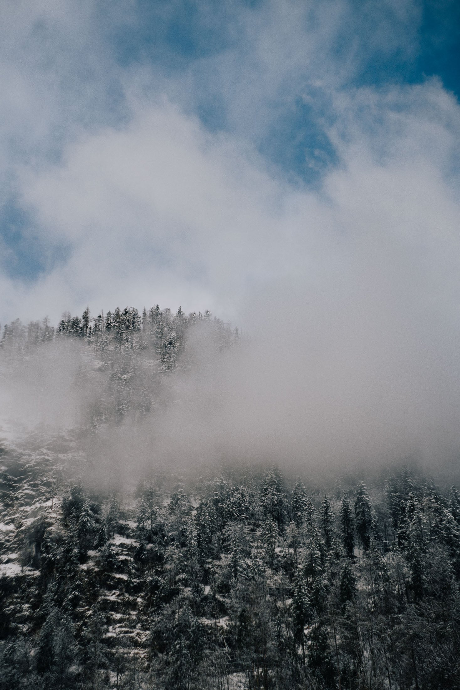 A snowy mountain landscape partially shrouded in mist and clouds under a blue sky.