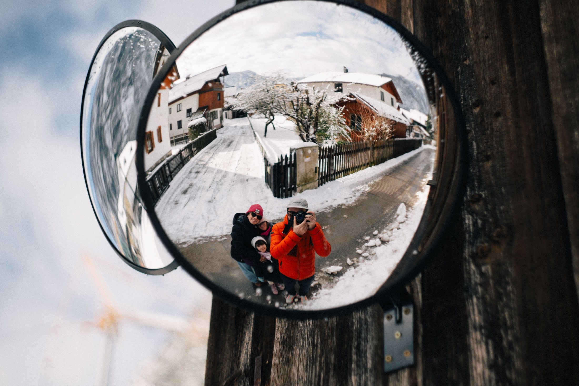 A reflection in a convex mirror showing a snowy street with wooden houses and a family taking a selfie, two adults and a child, bundled up in winter clothing.
