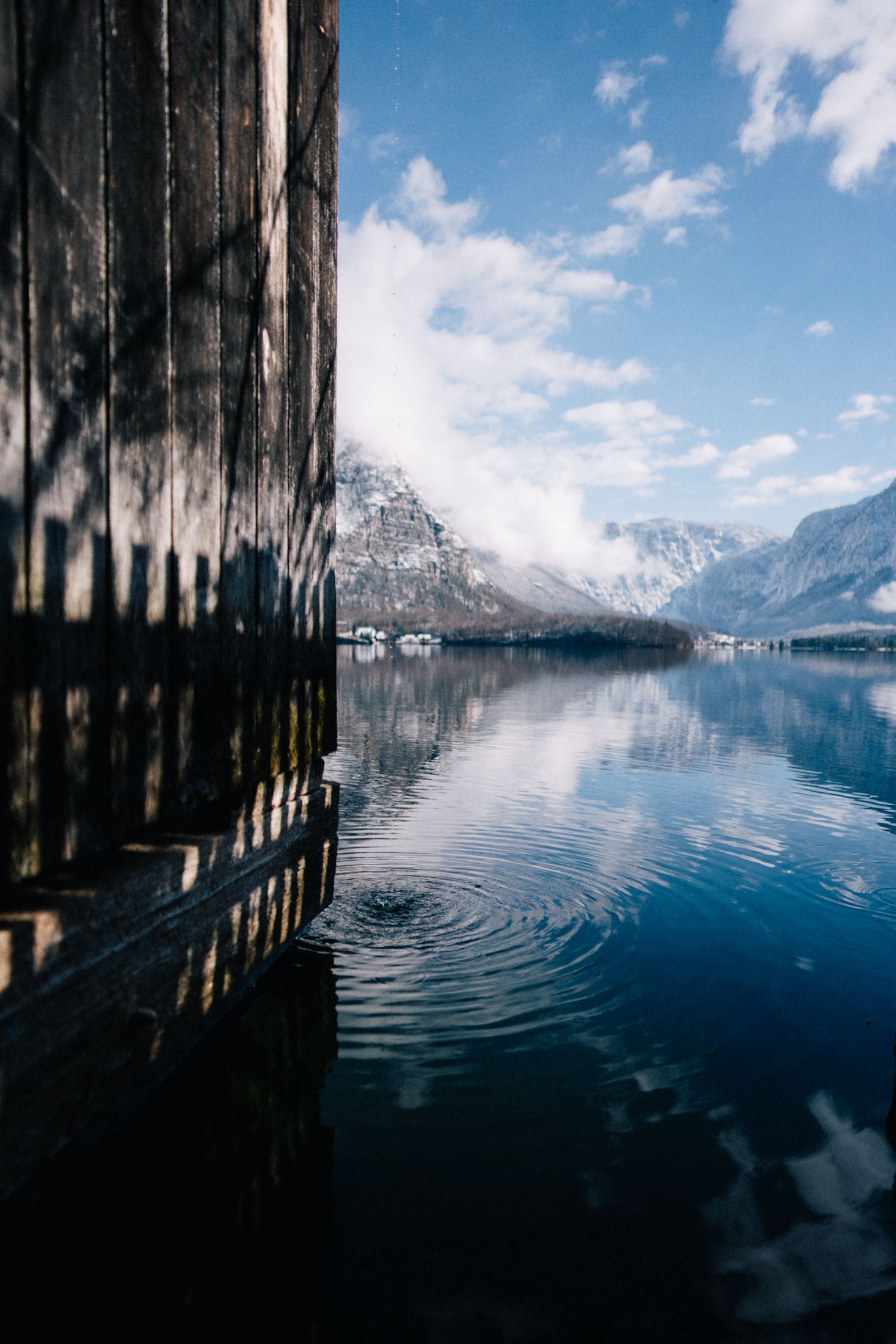 A serene lakeside view featuring a wooden structure on the left side, with reflections in the calm water and mountains in the background under a partly cloudy sky.