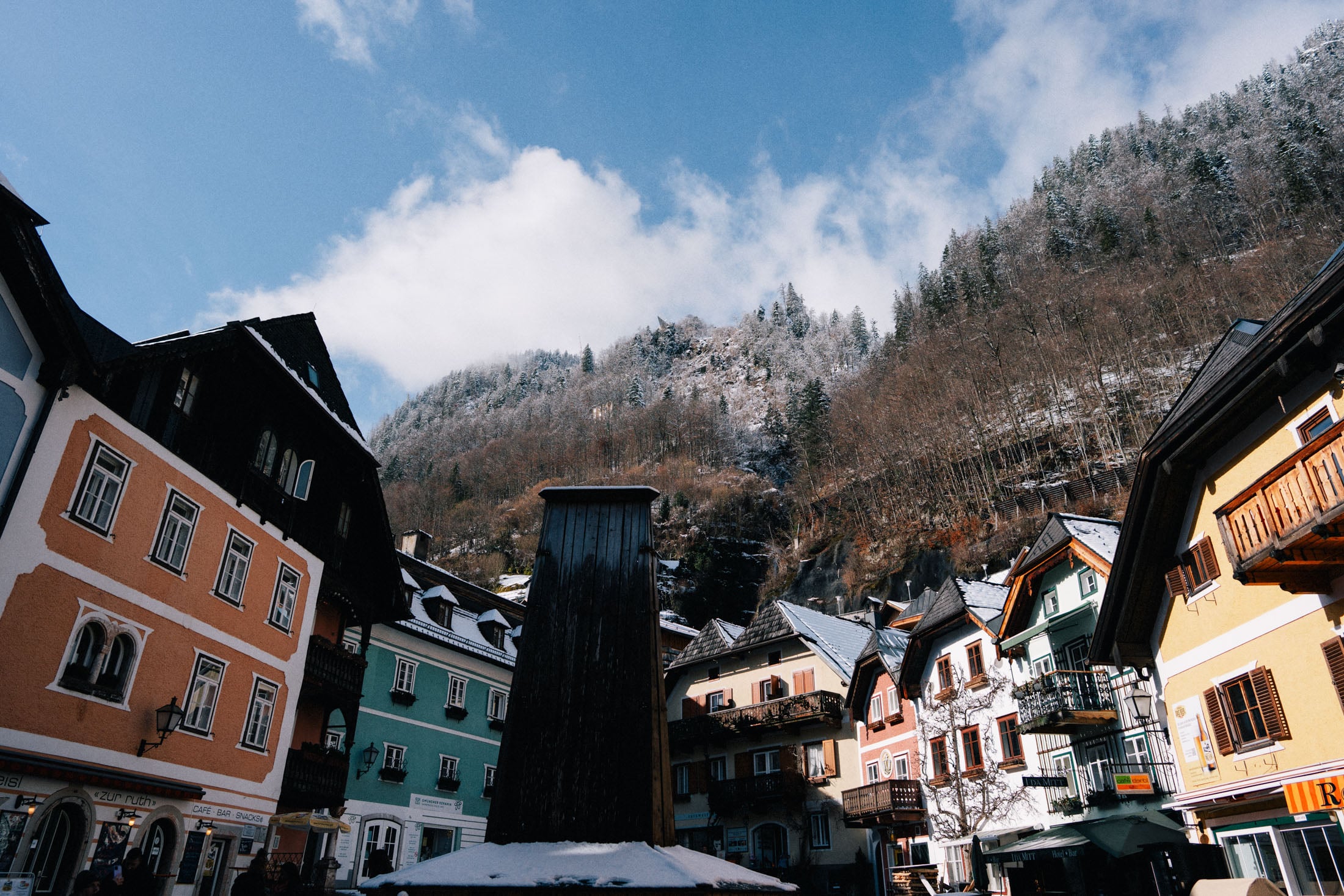 Charming alpine village with colorful houses, a central wooden structure, and snow-covered mountains in the background under a partly cloudy sky.
