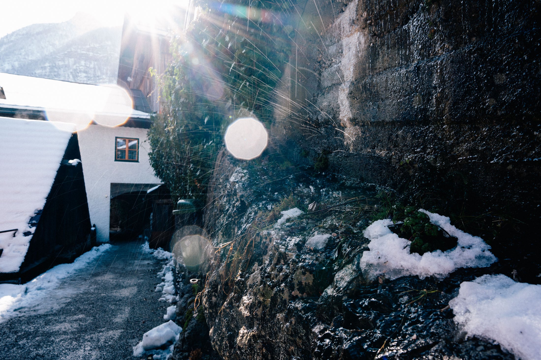 Sunlight shines down an icy pathway between buildings, with snow and patches of grass visible along the rocky wall.