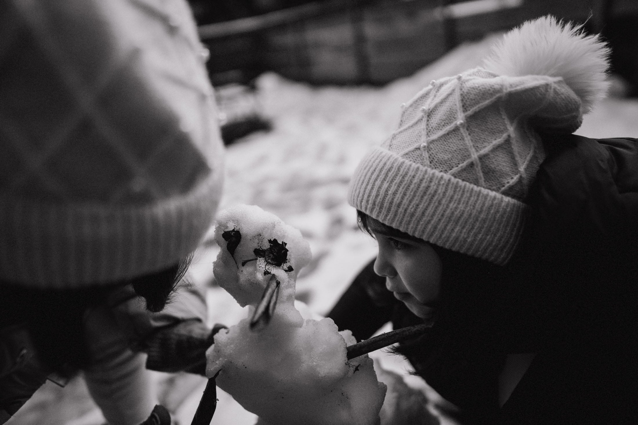A child in a light knit hat closely examines a snowman made of snow, decorated with sticks and leaves, in a snowy outdoor environment.