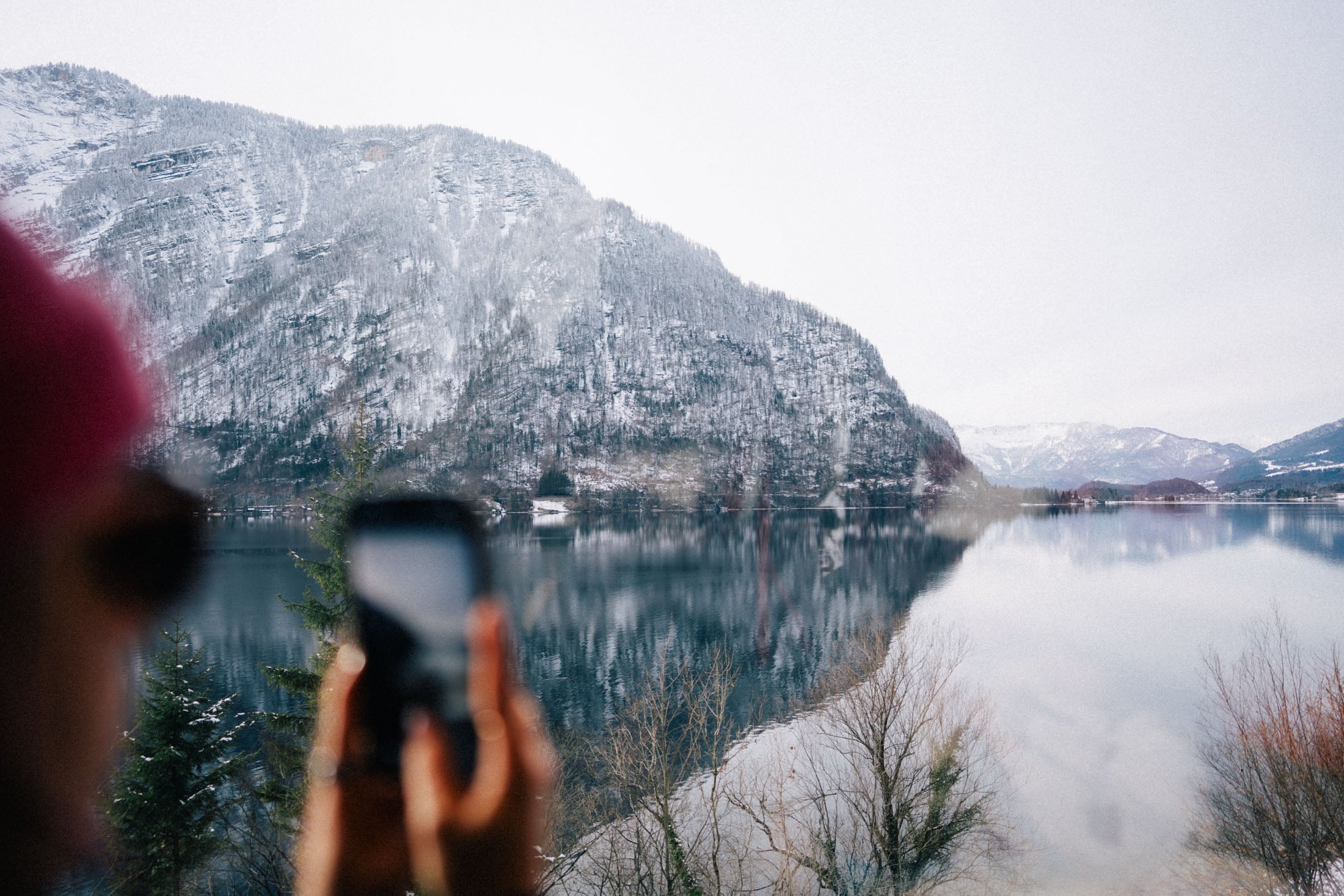 A person holding a smartphone in front of a picturesque snowy mountain landscape reflected in a calm lake.