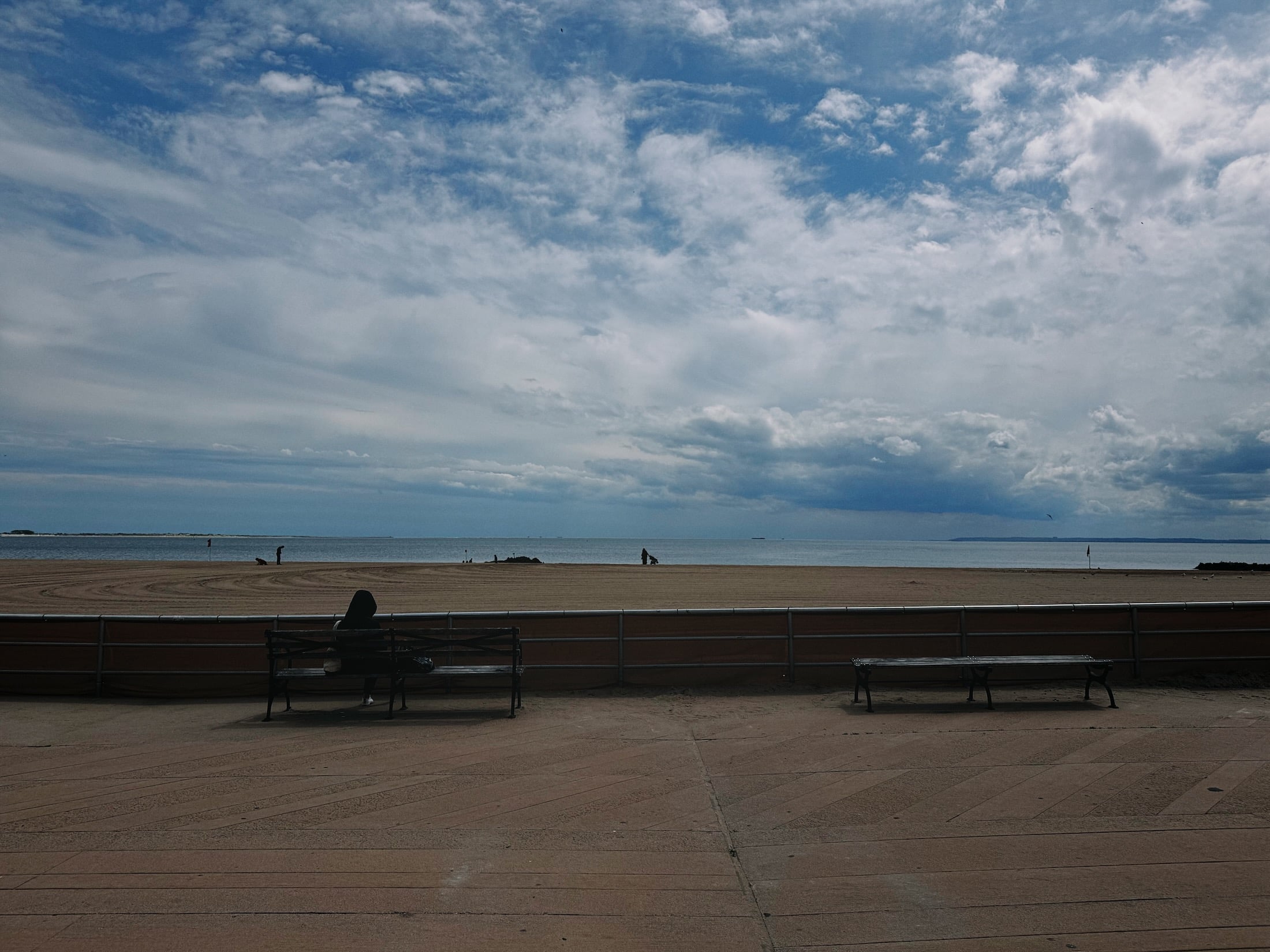 A serene beach scene with a sandy shore, gentle waves, and a cloudy sky. Benches are visible in the foreground, and a lone figure is walking along the beach.