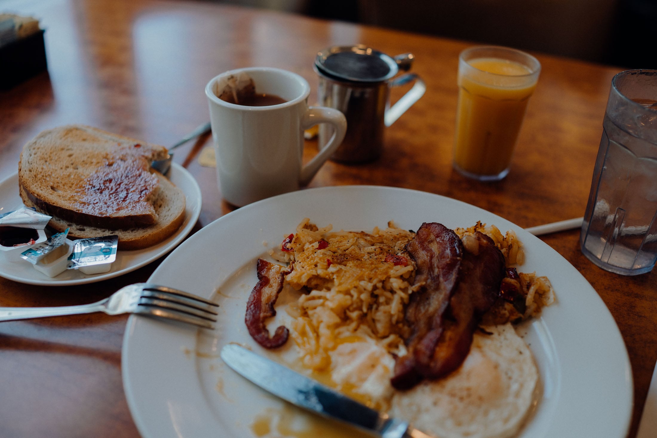 A breakfast plate featuring scrambled eggs, crispy bacon, and hash browns, alongside a slice of toast with jam and assorted butter packets. A cup of coffee and a glass of orange juice are also on the table.