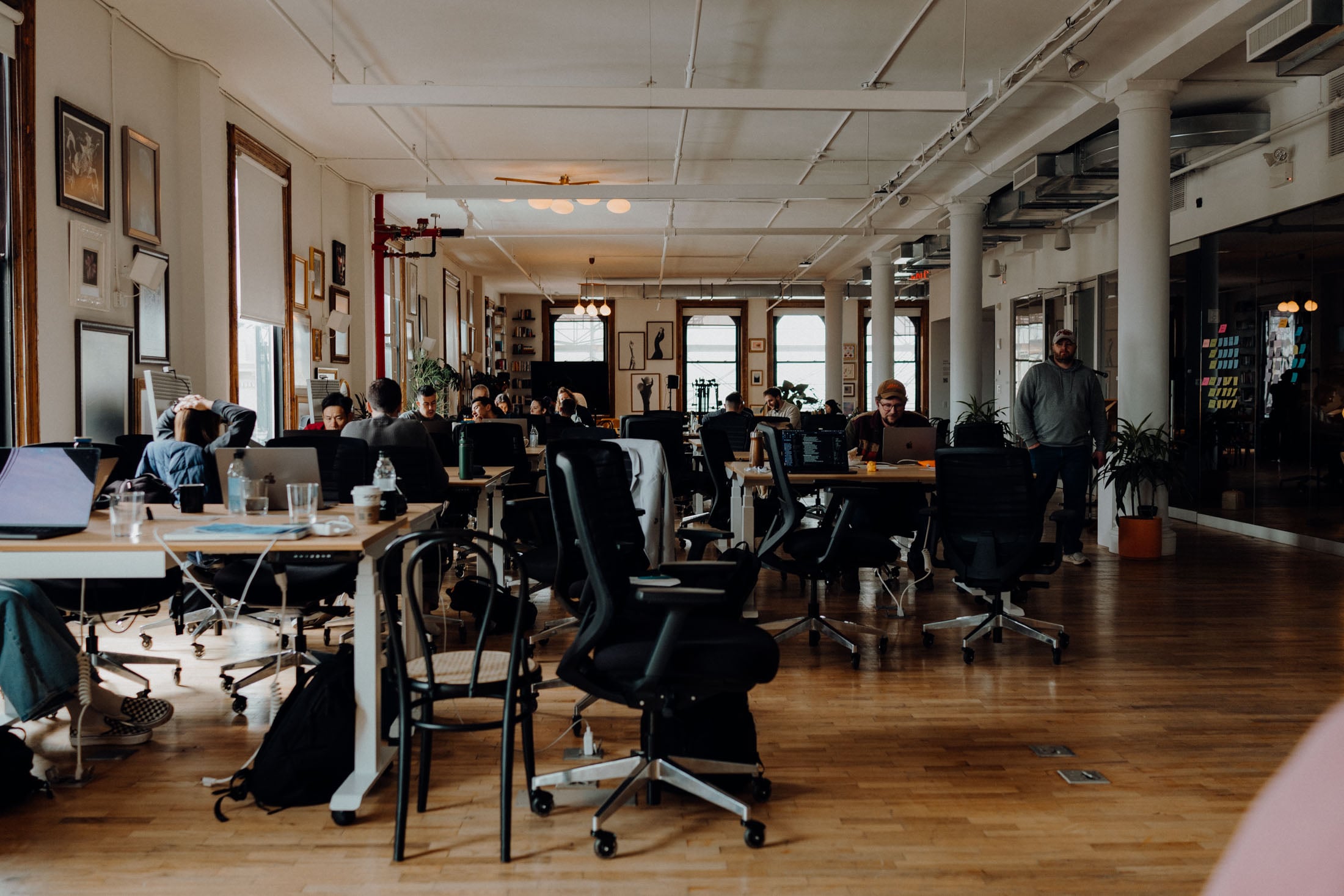 A modern coworking space filled with people working at desks with laptops. The environment features large windows, wooden flooring, and decorative wall art.