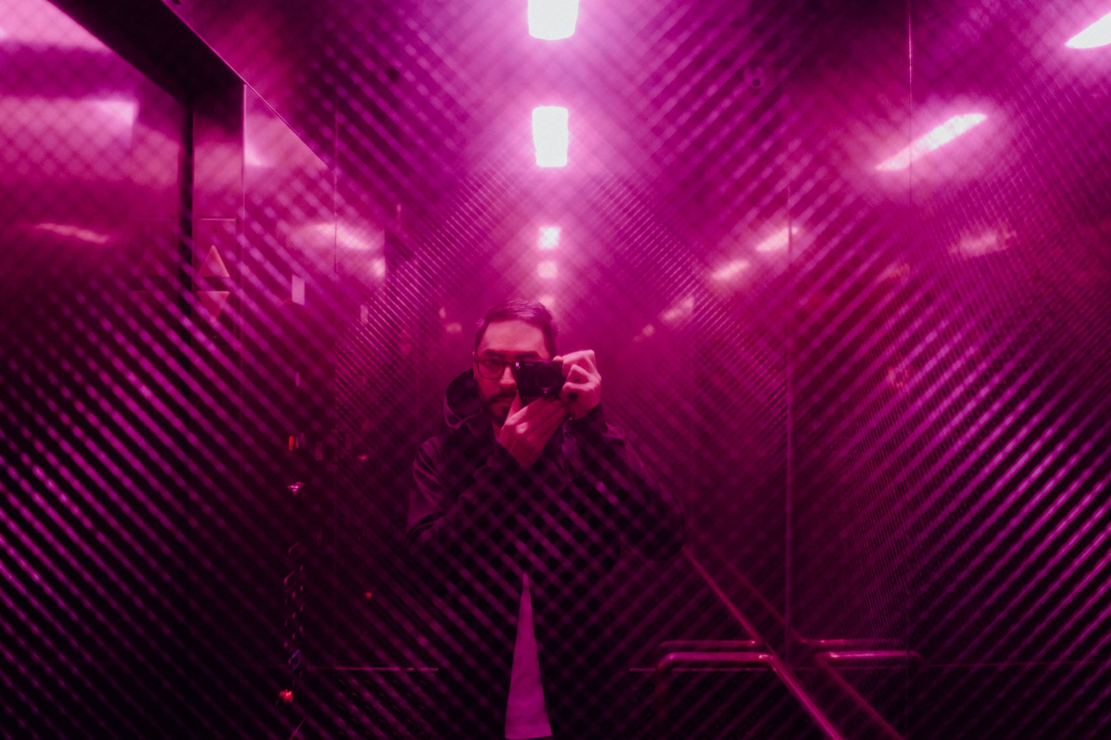 A person holding a camera is reflected in a purple-tinted elevator, surrounded by shiny surfaces and bright lights.