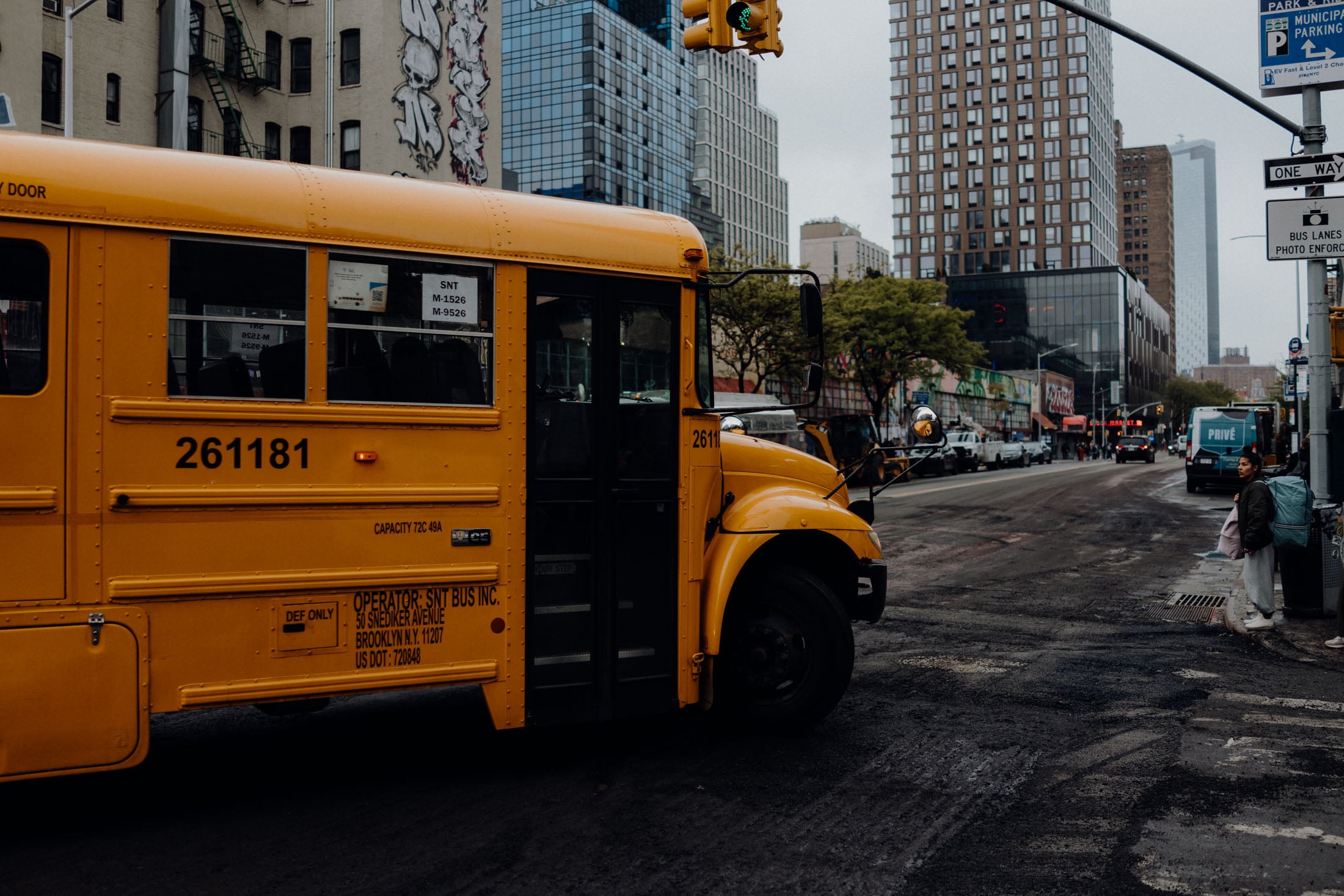 A yellow school bus parked on a city street with buildings in the background. The street shows signs of recent work, and a person stands on the sidewalk nearby.
