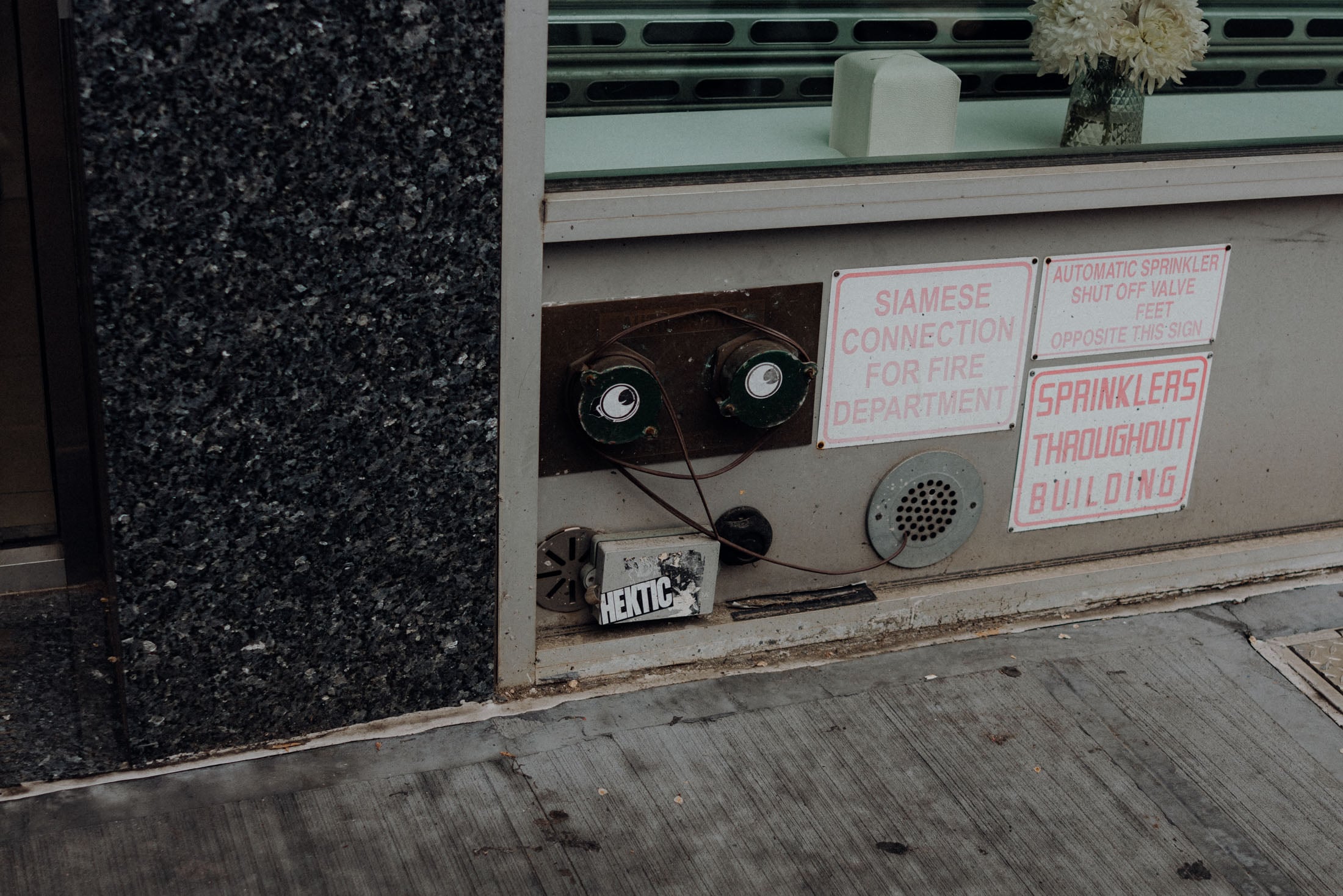 A close-up view of a fire department sprinkler connection mounted on a wall, featuring green valves, several informational signs about fire safety, and a box labeled 'HEKTIC' on the ground.