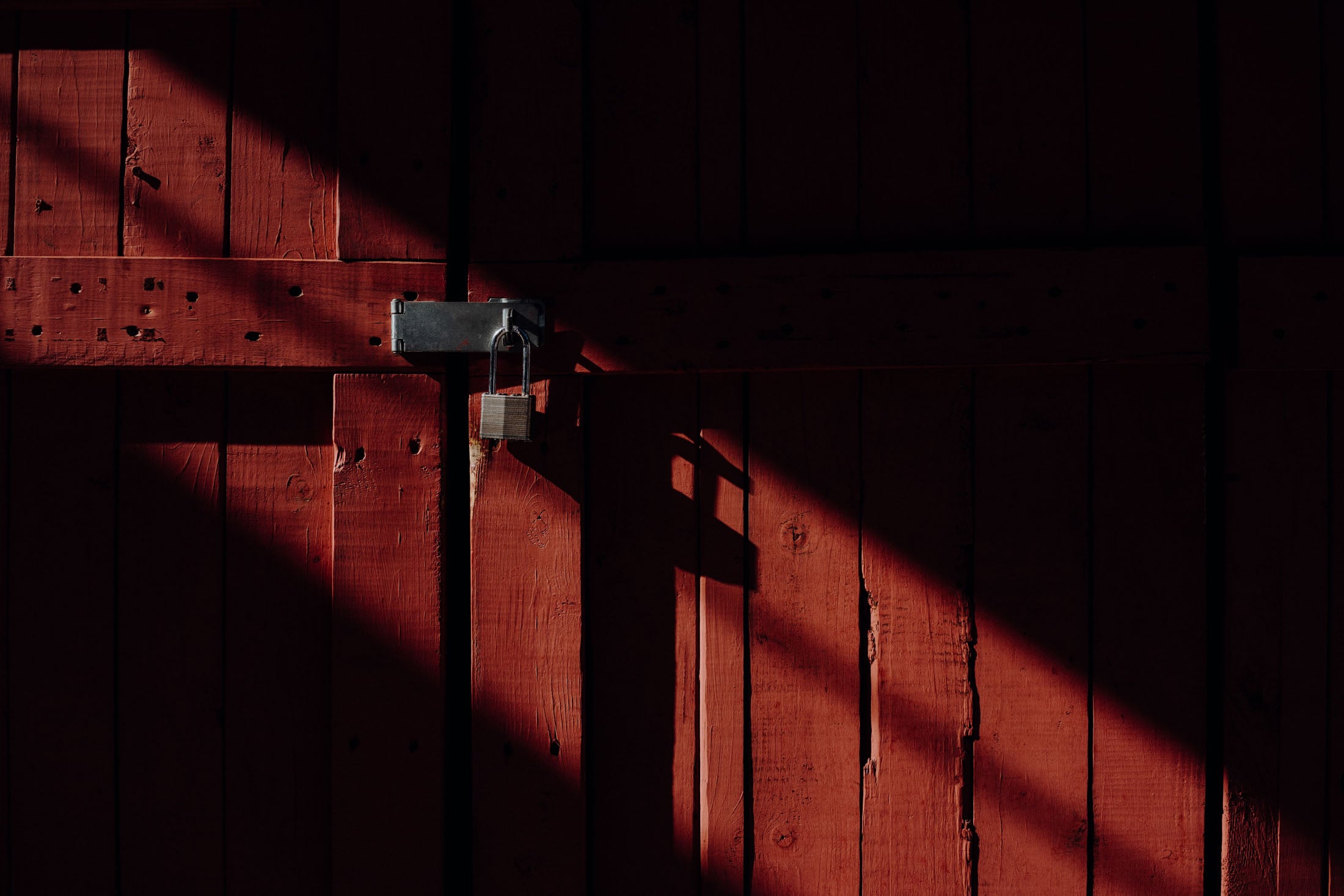 Close-up of a red wooden door with a padlock, featuring shadows and sunlight casting diagonal patterns.